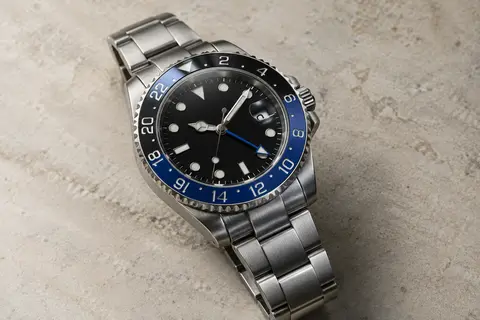 Montre GMT automatique à cadran noir, lunette céramique noire et bleue, boîtier et bracelet en acier sur fond minéral ivoire.