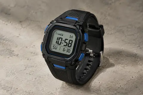 Montre digitale noire à bracelet en silicone, écran LCD et détails bleus, posée sur un fond minéral clair.
