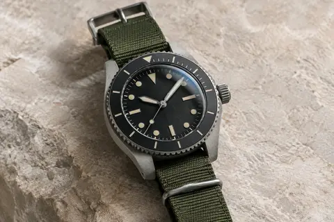 Montre de terrain Timex à cadran noir, boîtier gris et bracelet textile vert, photographiée sur fond minéral.